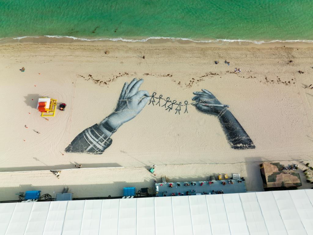 Sand art of two hands holding a rope of stick figures holding hands on a beach in Miami