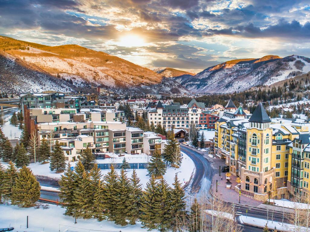 Panoramic view of colourful buildings lit by golden sunlight through clouds, surrounded by mountains with snow and pine trees