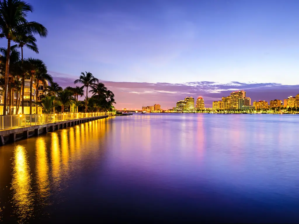 West Palm Beach, Florida with at night with the city lights reflecting in the water in the foreground and tall palm trees lining the waters edge.