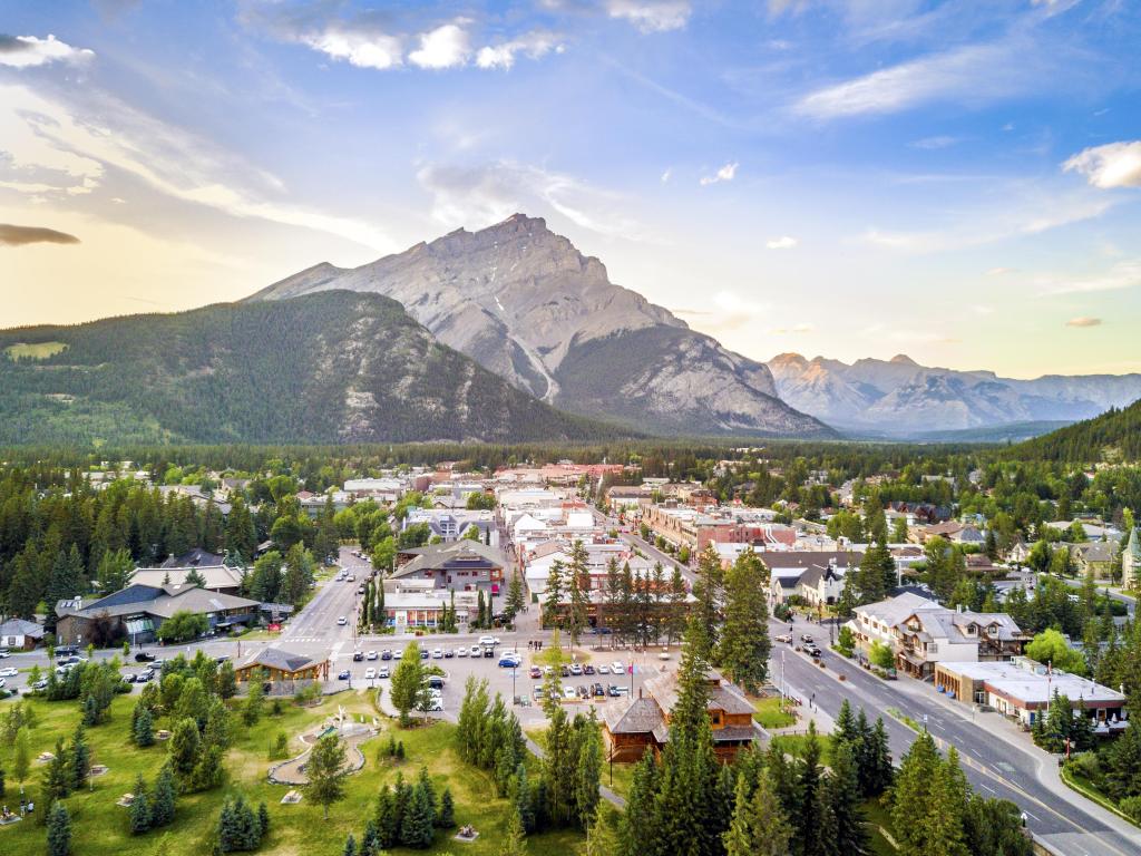 Banff, Rocky Mountains, Alberta, Canada with the city in the foreground, dotted by trees and the rocky mountains in the distance on a sunny clear day.