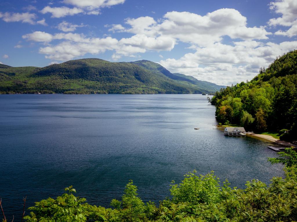 Lake George, New York, USA with a view of the bay surrounded by trees and green hills in the distance on a sunny and cloudy day.