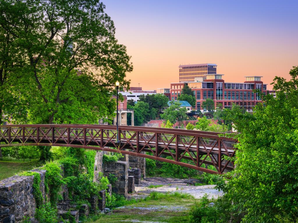 Columbus, Georgia, USA downtown skyline and park with a bridge crossing over a river and lots of trees and greenery in the foreground at sunset.
