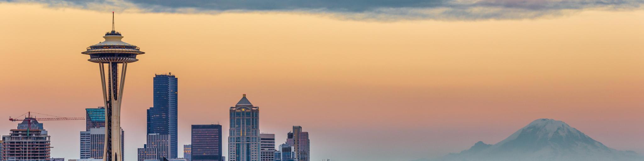 Seattle Skyline at sunset. The photo is taken on a slightly cloudy day and the clouds reflect the dramatic colors of the sunset. Space Needle can be seen.