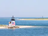 The Nantucket harbor lighthouse amidst a beautiful sunny day while on the upper right of the image is a sailboat.