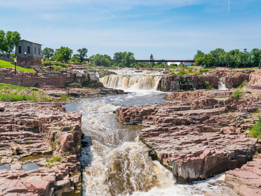 Falls Park, Sioux Falls, South Dakota with the Big Sioux River and small waterfalls running down alongside the rocky terrain, trees and a bridge in the distance on a sunny day.