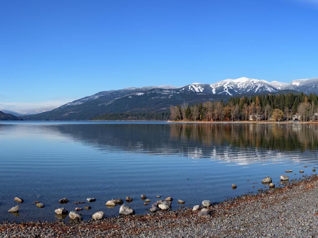 Whitefish Lake, Montana with a panorama shot of the lake and snow-capped mountains in the distance, stony shore in the foreground and on a sunny day with a pure blue sky.