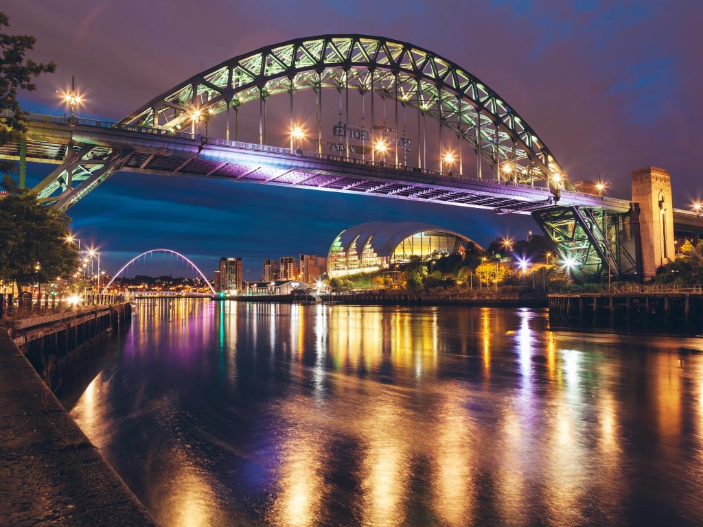 The Tyne Bridge, Newcastle, UK with a view of the river and bridge at night.