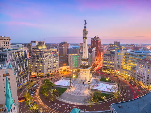 Indianapolis city skyline at sunset with a purple hued sky behind and Soliders' and Sailors' Monument below