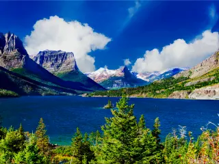 Stunning view of St. Mary Lake surrounded with tall green trees and the mountains on a sunny day with blue skies and fluffy white clouds.