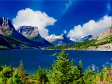 Stunning view of St. Mary Lake surrounded with tall green trees and the mountains on a sunny day with blue skies and fluffy white clouds.