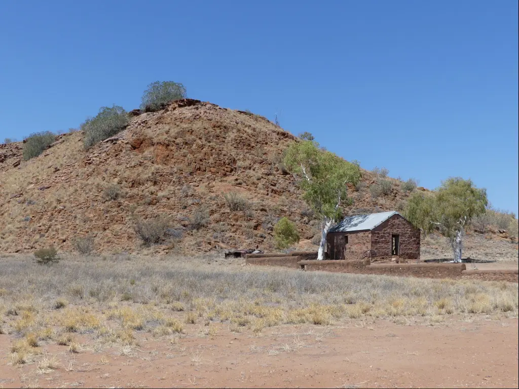 Lonely, old sandstone brick house between two white ghost gum trees by a hill at the old Barrow Creek Telegraph Station in Northern Territory, Australia