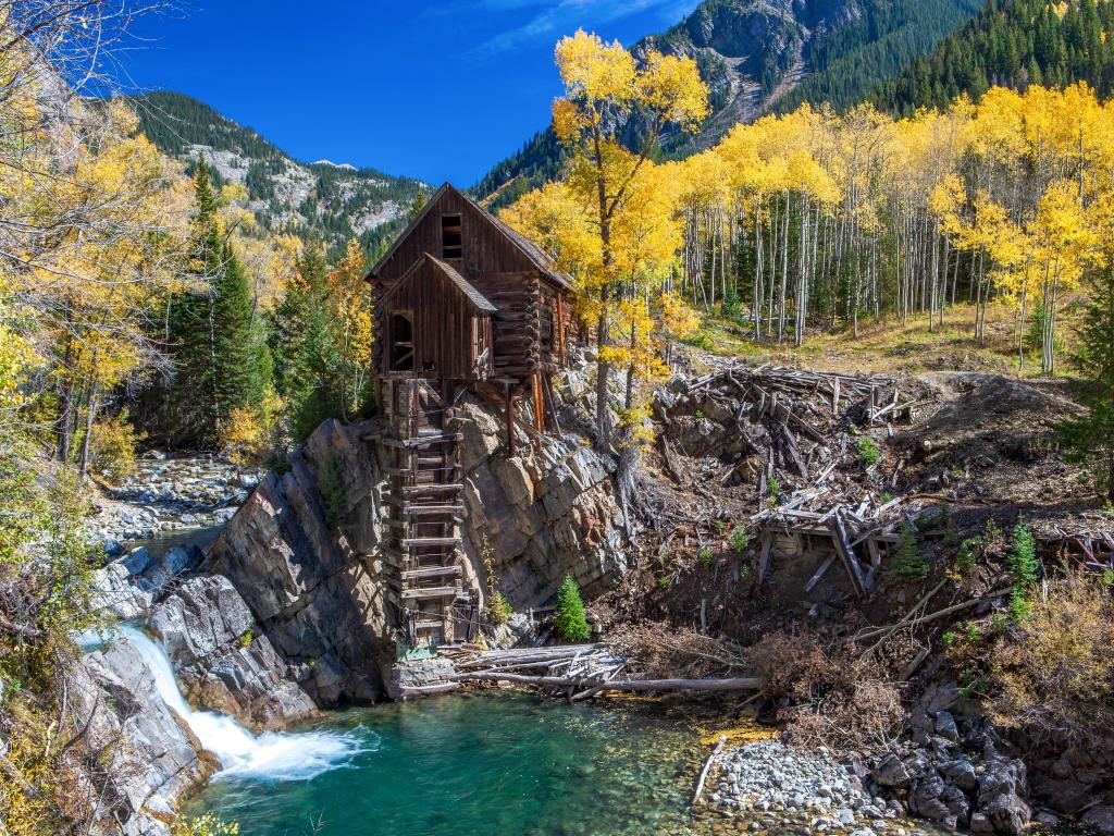 Log-built mill building perched on a small rocky cliff above a pool, with blue sky and forested mountain slopes in the background