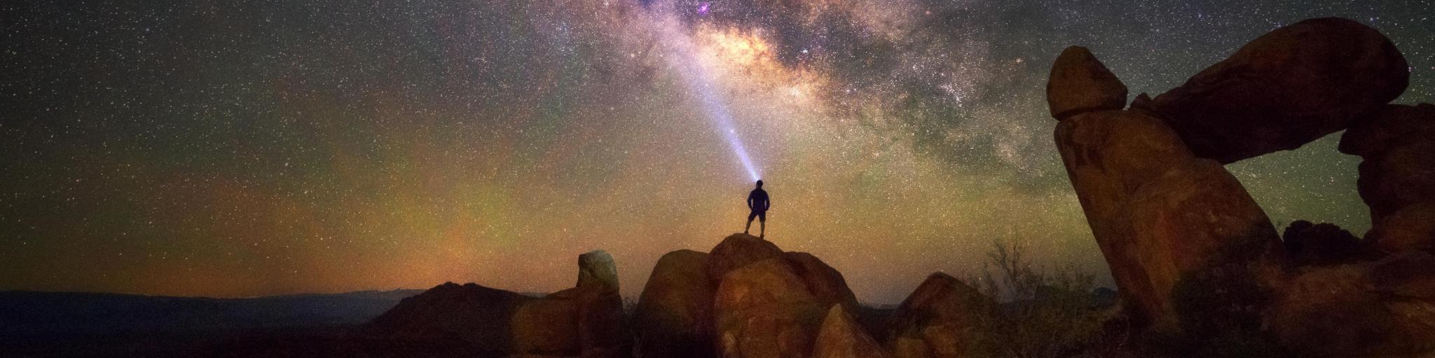 Night time view of star constellation and galaxy at Balanced Rock, Big Bend National Park, with silhouetted profile of someone standing on the rockface