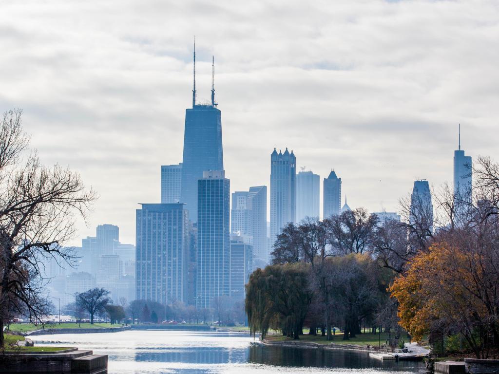 High rise buildings appear grey in the mist behind a calm lagoon in the foreground with autumn trees