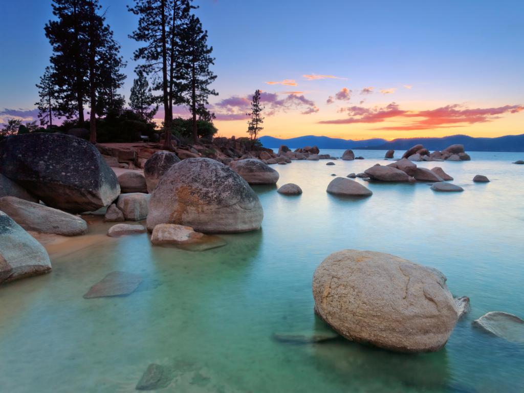 Orange and pink sunset light reflected into still turquoise water by a lake shore with large boulders
