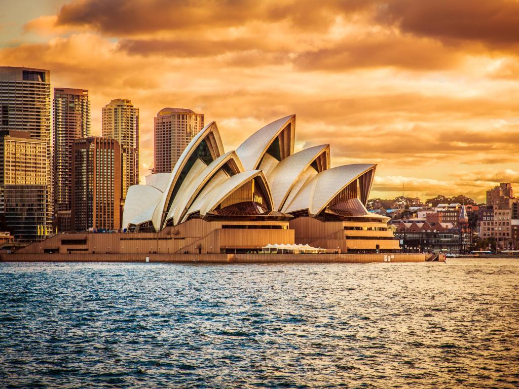 Sail-shaped outline of Sydney Opera House viewed from a ferry travelling across the harbour, lit up in golden light