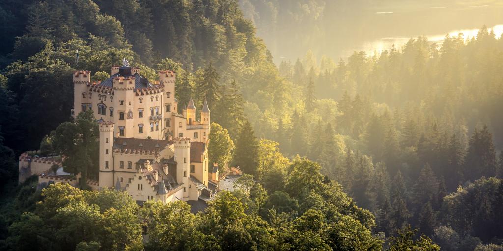 Hohenschwangau Castle in Fussen, Bavaria, peeking out of the surrounding forest, with a ray of sunlight hitting it