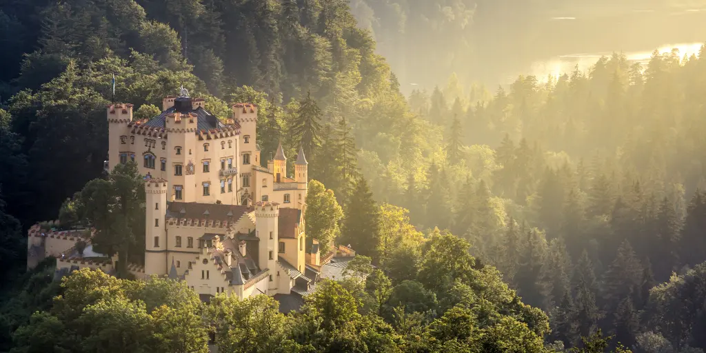 Hohenschwangau Castle in Fussen, Bavaria, peeking out of the surrounding forest, with a ray of sunlight hitting it