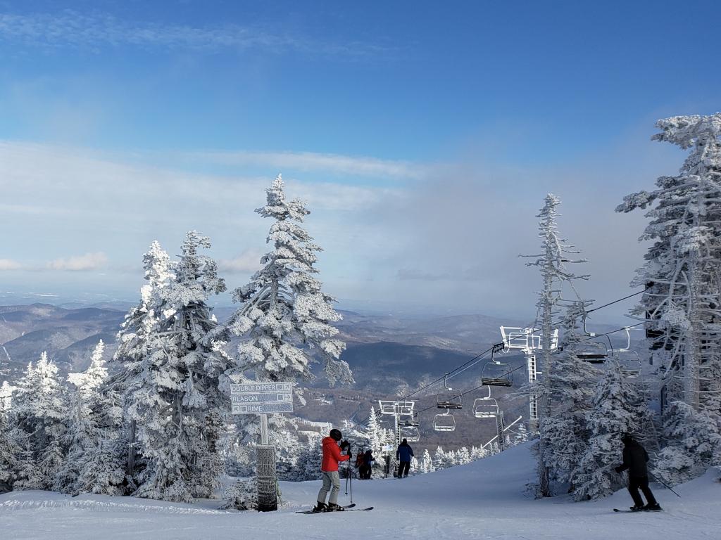 Two skiiers standing at the top of a piste in Killington, Vermont, surrounded by snowy trees and with a chair lift travelling overhead