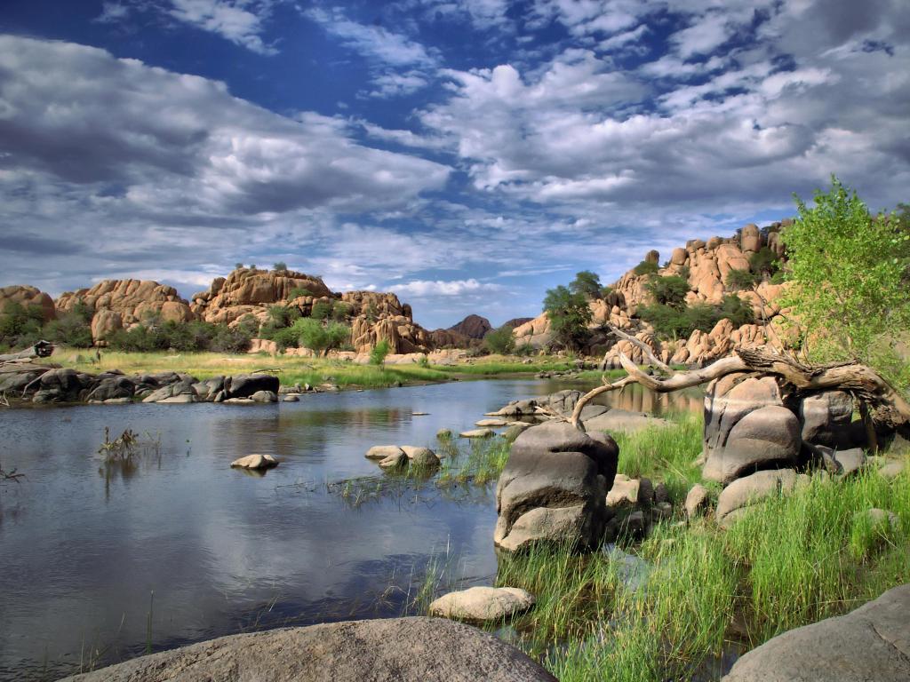 A view from the shore of Watson Lake Park, Arizona, USA with blue cloud filled sky and green trees with rock cliffs.