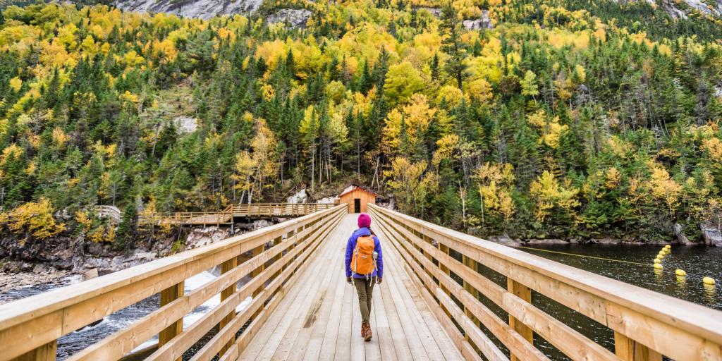 A women walking over a wooden bridge towards a hill covered in trees in a national park in Quebec 
