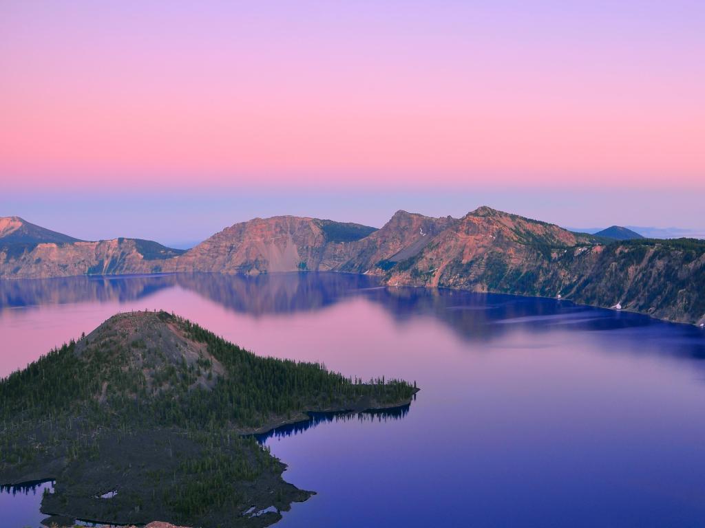 Pink sky over calm blue lake with part of steep-sided crater rim in view