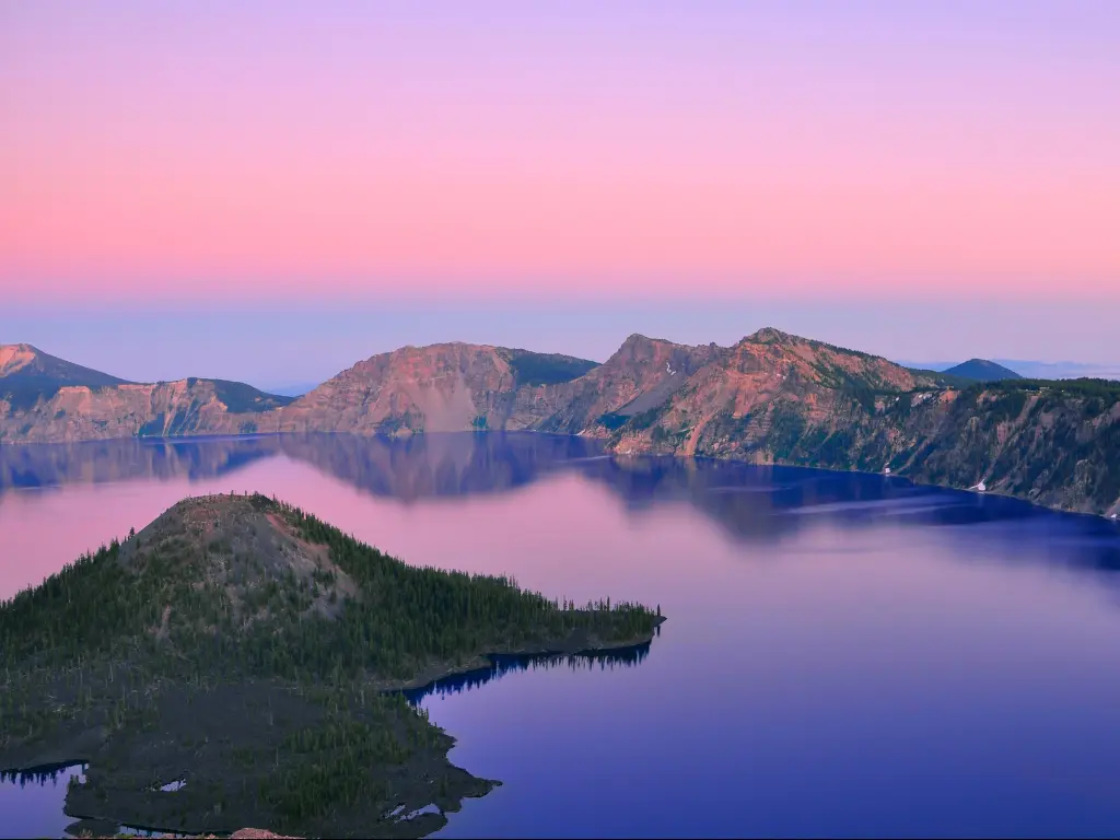 Pink sky over calm blue lake with part of steep-sided crater rim in view