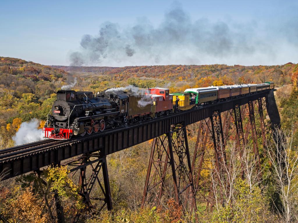 Steam train traveling on a railway bridge with scenic views of autumnal trees below