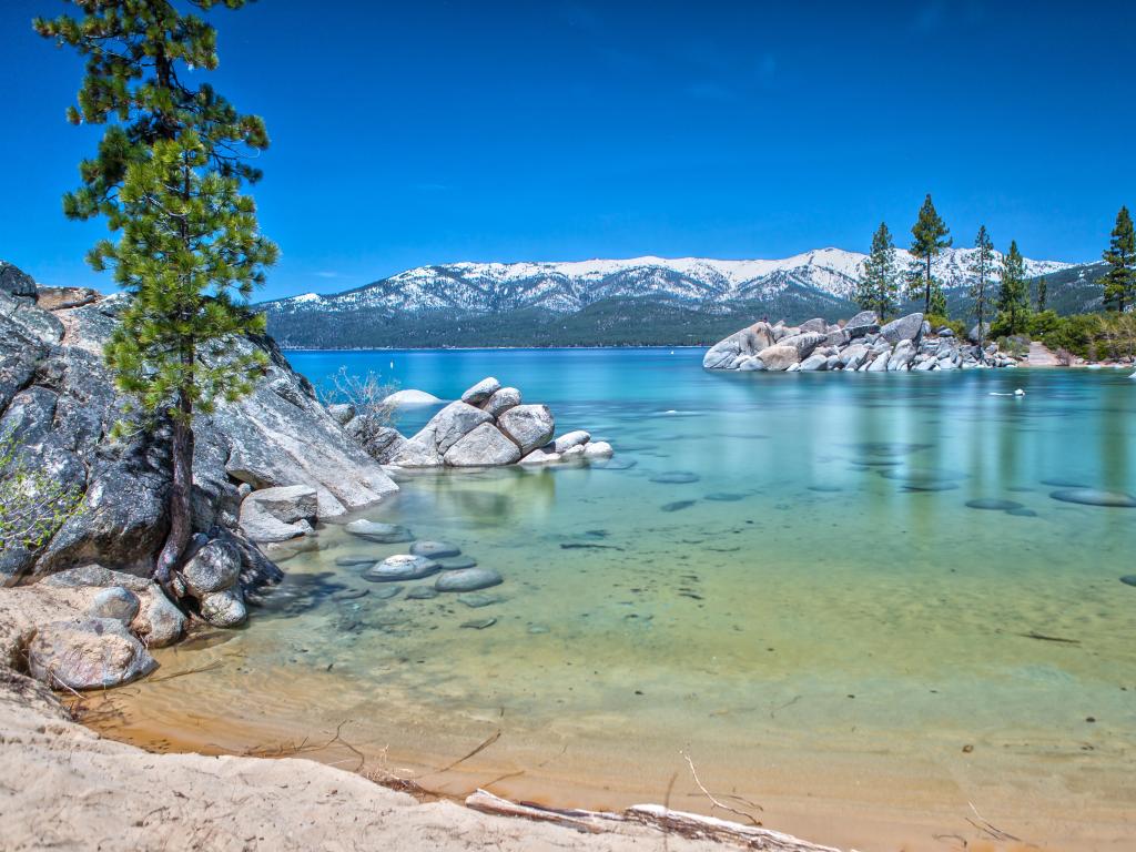 View of shallow, clear water on the edge of a wide lake with snow-capped mountains on the opposite shore and blue sky