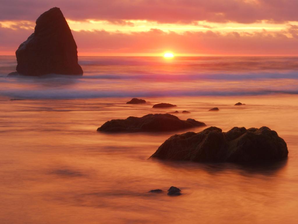 Rock rising out of calm ocean by beach lit up in vibrant gold and pink sunset light