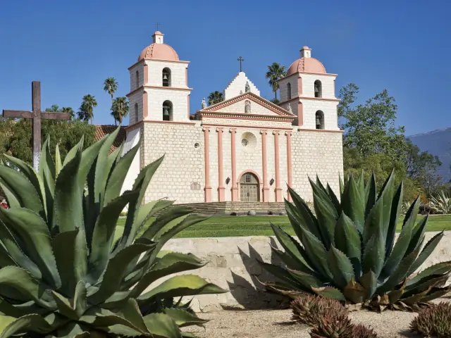 Pink and white mission building on a sunny day with aloe vera plants in the foreground