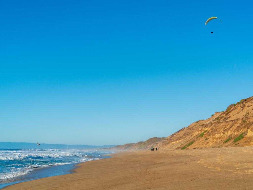 Sandy beach with turbulent waves, several people walking in the distance with a person paragliding