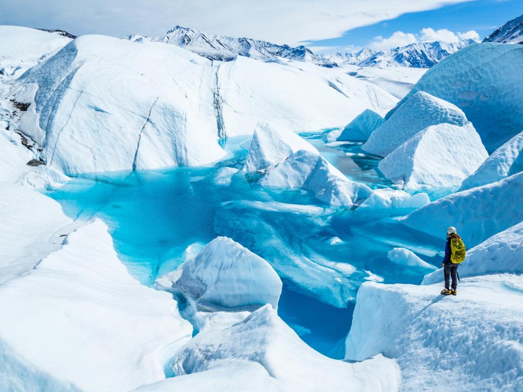 Standing near the edge of large blue pool on top of the Matanuska Glacier. A young woman holding an ice axe with a backpack and helmet looks out over the lake.