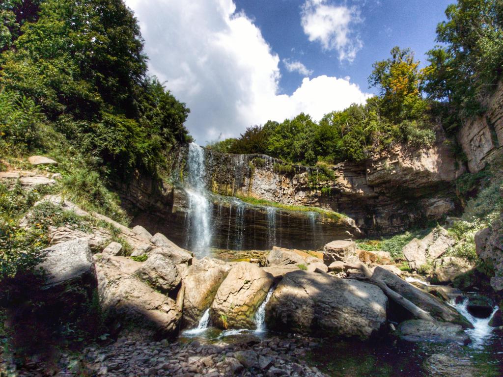 Webster Falls, Hamilton, Ontario, Canada taken on a sunny day with the waterfall surrounded by green foliage and landing on the rocks below.