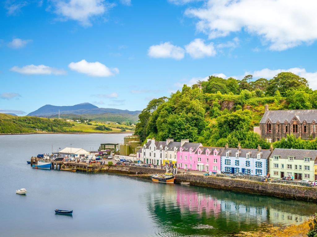 Portree harbor, Isle of Skye, UK with colourful buildings lining the shore and reflecting in the water, against greenery and a blue sky.