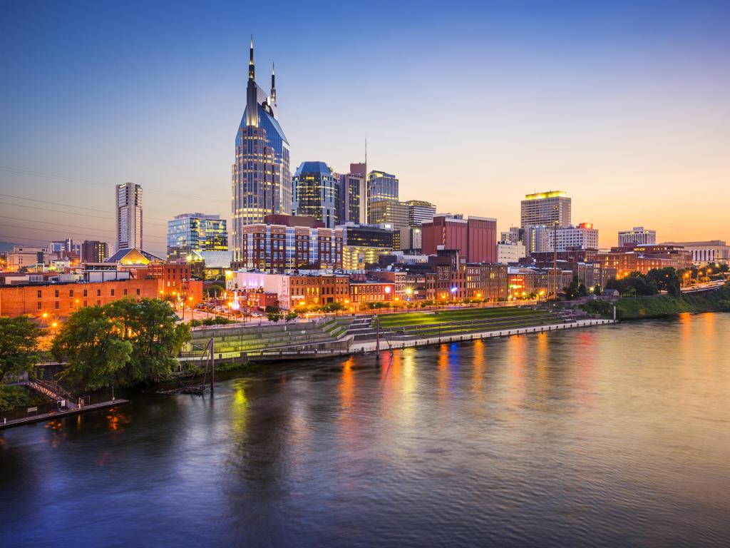 Nashville, Tennessee at sunset with the city skyline lit up and overlooking the river in the foreground. 