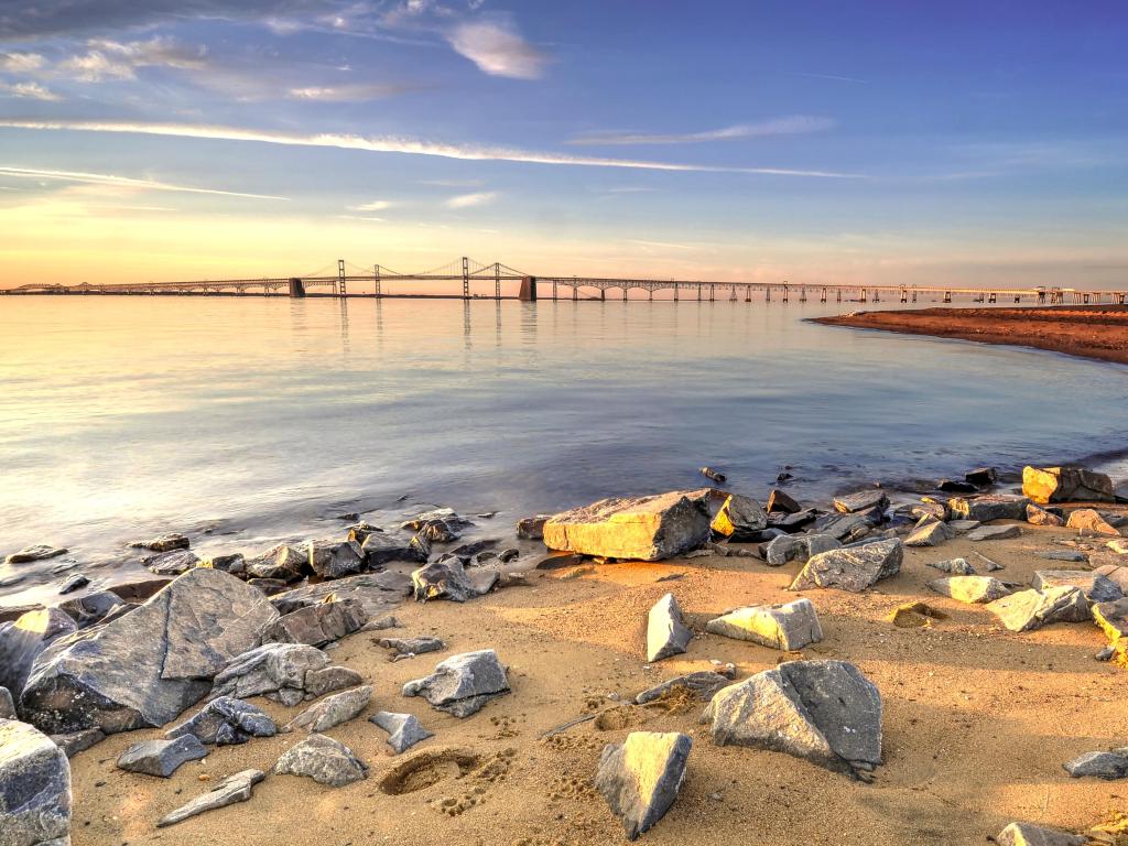 Chesapeake Bay Bridge, Chesapeake Bay, USA taken at sunrise with a colorful morning sky over the Chesapeake Bay Bridge and the beach in the foreground with an assortment of stones lie peacefully, soaking in golden sunlight.