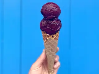 An ice cream cone with a berry-flavored ice cream is held up in front of a blue painted wooden shiplap wall