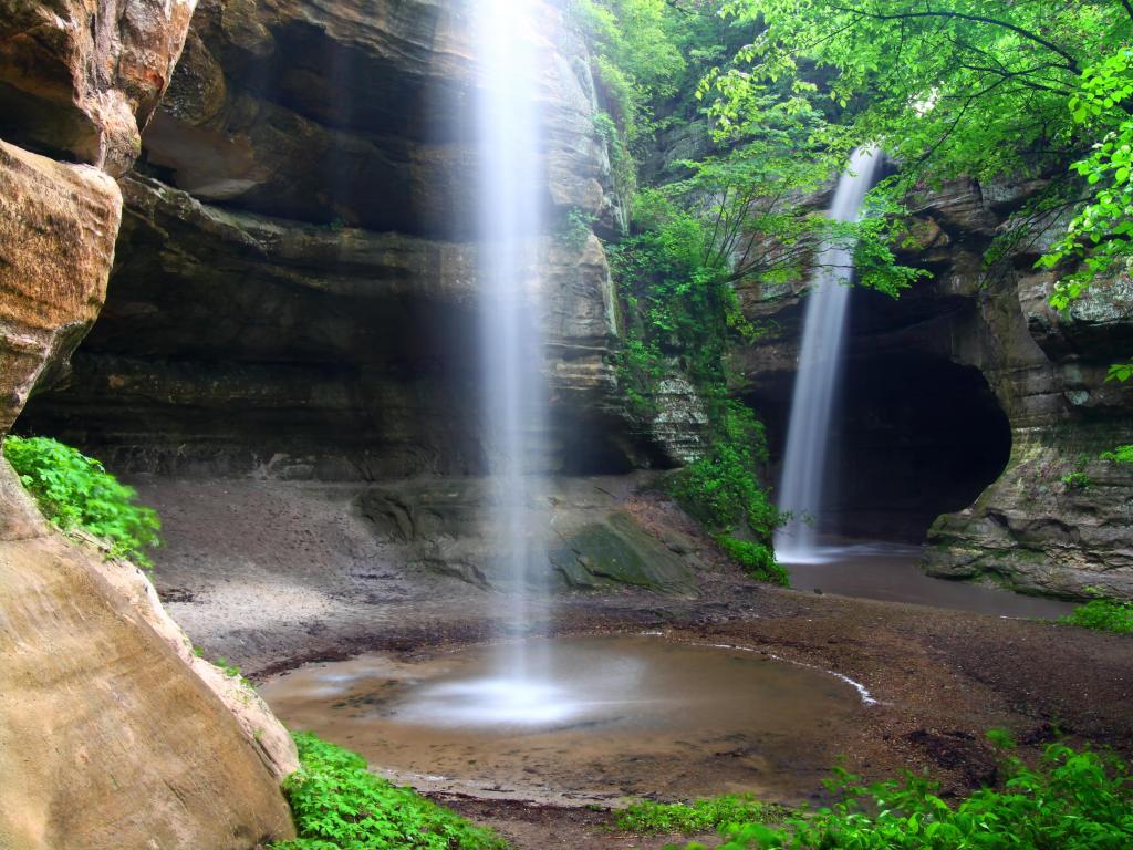 Starved Rock State Park, Illinois, USA with a view of twin waterfalls crashing into Tonti Canyon on a spring day surrounded by green trees.