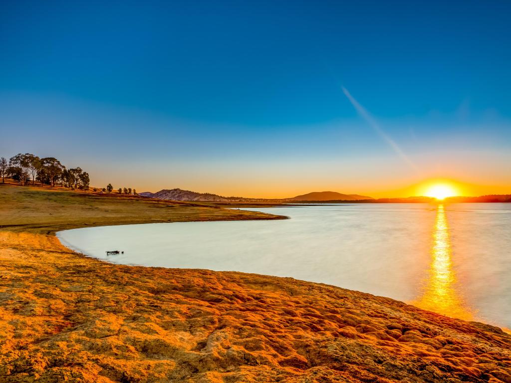 Sun rising over still water of Lake Hume with vivid reflection and a few gum trees on the banks