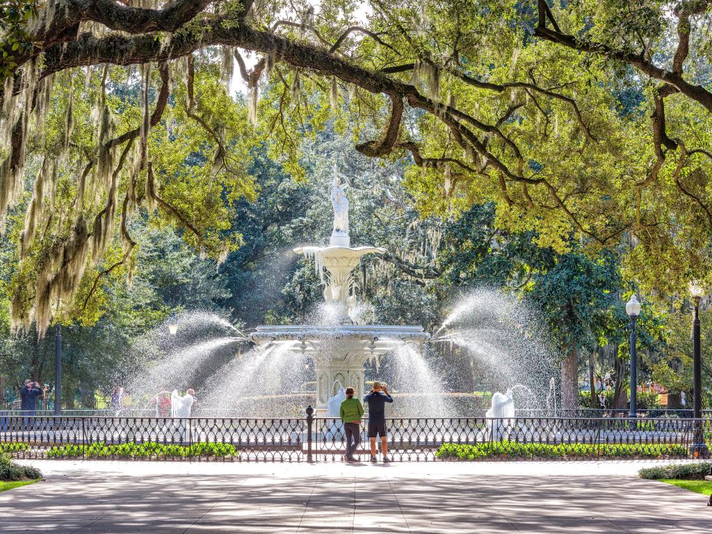 Tourists seen admiring the fountains in Forsyth Park, Savannah on a sunny day, viewed through the branches of the park's southern oaks