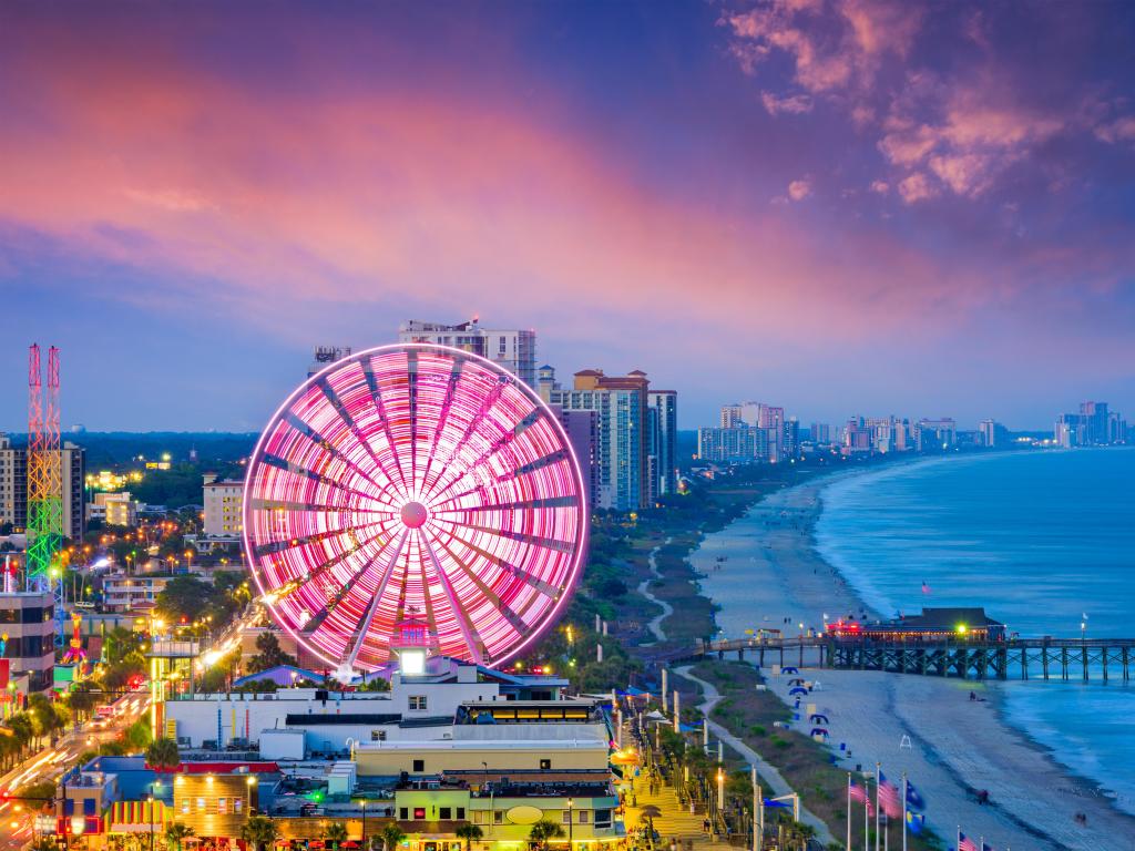 Ferris wheel by the beach lit up pink, with a line of high rise buildings stretching out into the distance along the waterfront