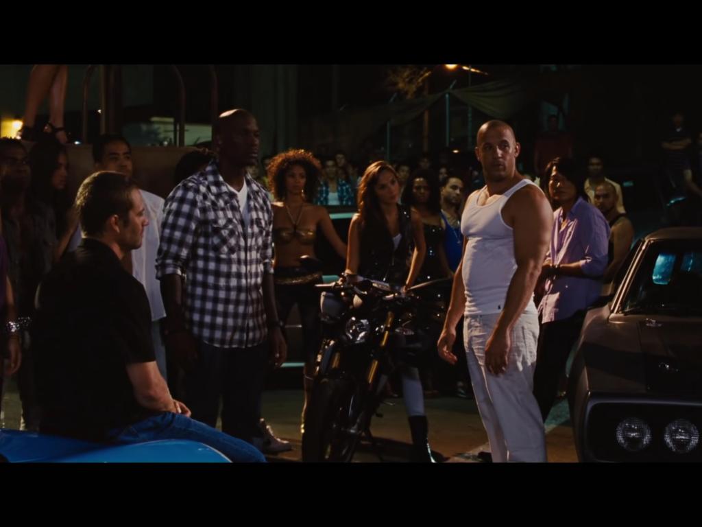 Standing between a motorbike and a car, Dom is surrounded by his team and his friends as he looks to his left. This scene in Fast Five takes place at night.