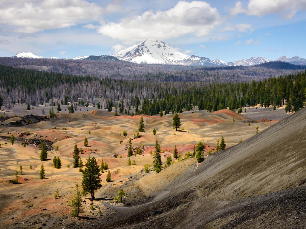 Lassen Volcanic National Park, California with a sandy terrain in the foreground, dotted with trees and a forest and snow-capped mountain in the background.