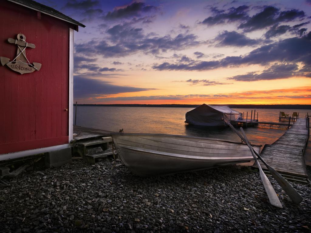Sun sets over a calm lake. A small pier reaches out from a stony beach and a red storage shed on the beach has a wood anchor sign that says Welcome.