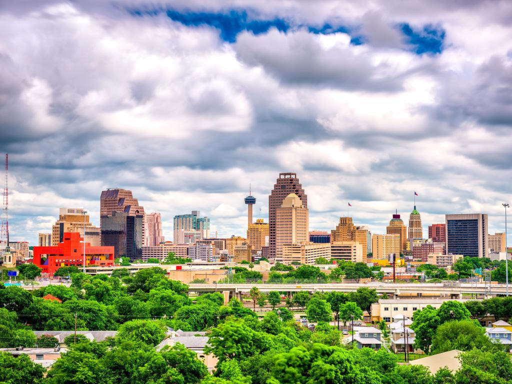 San Antonio, Texas, USA with trees in the foreground and the downtown cityscape in the distance against a cloudy sky.