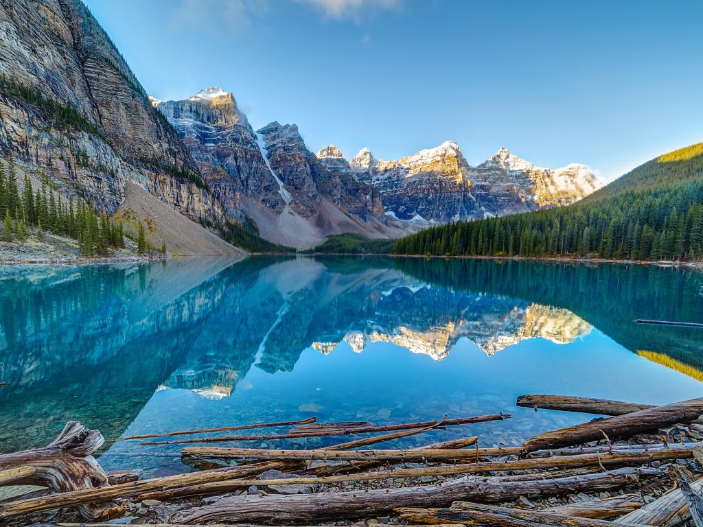 Turquoise lake reflects snow-covered mountain peaks with clear blue sky and sloping, tree-covered lake shores