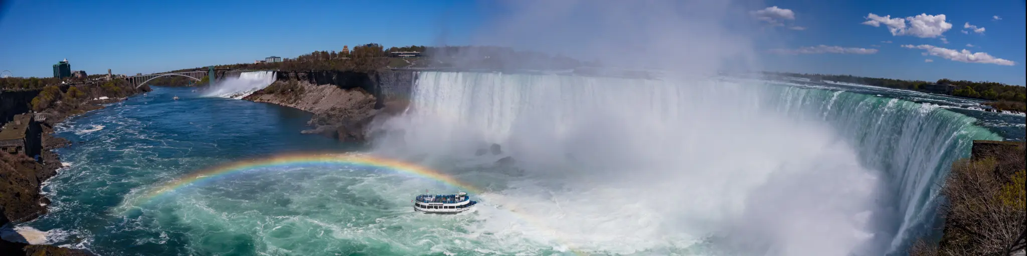 A panoramic view of Niagara Falls with a boat near the mist from the water that creates a rainbow.