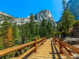 View of Half Dome and Liberty Cap peaks of Nevada Fall in the distance over a wooden bridge on a sunny day