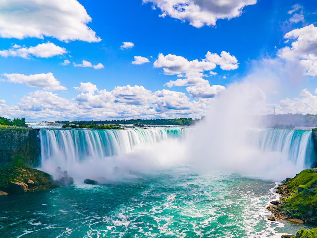 Niagara Falls, border of New York State and Canada, on a sunny day with blue sky, showing the incredible vast waterfall and water spray below. 